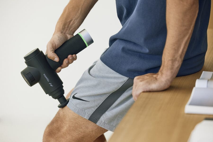 Personal trainer using a Hyperice massage gun as part of assisted stretching exercises to prepare the quadriceps before stretching.